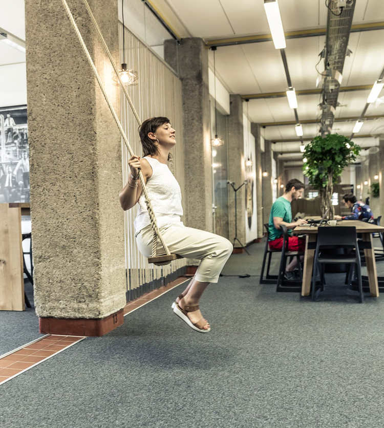 Eine Frau sitzt auf einer schwingenden Schaukel in einem modernen Innenraum. Ihr Gesichtsausdruck zeigt Freude und Gelassenheit. Im Hintergrund sind einige Personen an Tischen mit Laptops beschäftigt, während die Atmosphäre freundlich und kreativ wirkt.